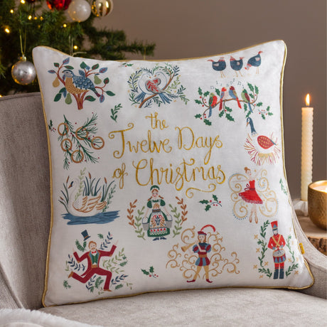 Cream velvet square cushion embroidered with "12 Days of Christmas" motifs in gold, multicoloured thread, and gold piped edge, shown on a sofa near a Christmas tree and candle.