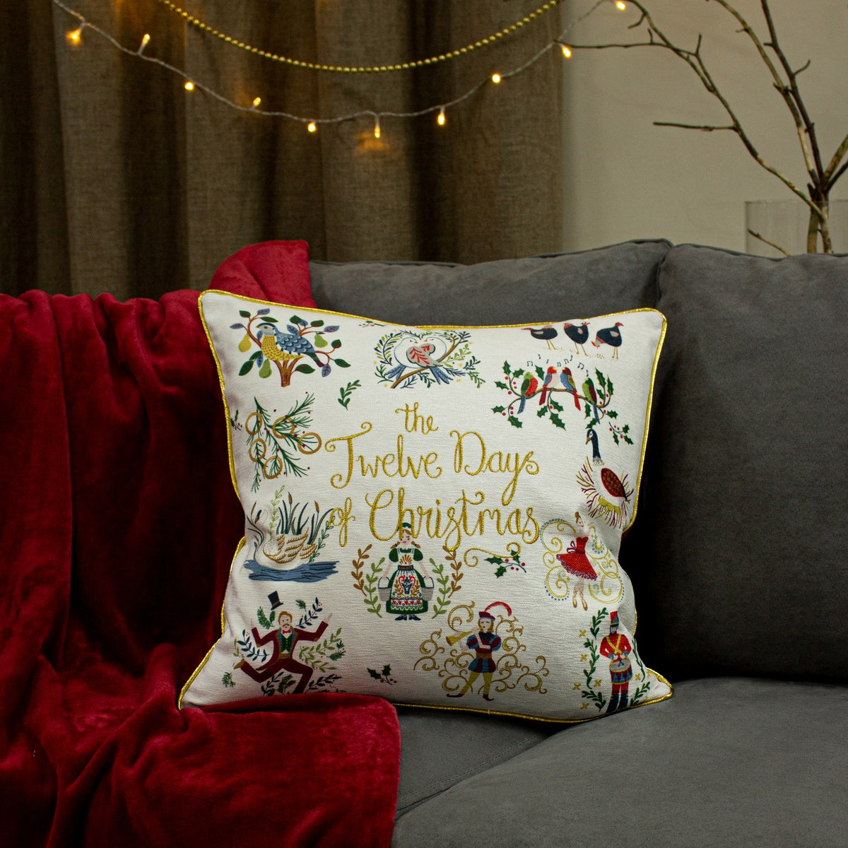 Cream square cushion with gold-piped edge, embroidered "Twelve Days of Christmas" motifs in multicolour, styled on a grey sofa with a red velvet throw.