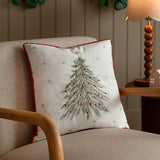 Twinkling Tree Velvet Linen Cushion in natural with red pom-pom trim, embroidered fir tree, gold stars and red baubles, styled on an armchair beside a wooden lamp.