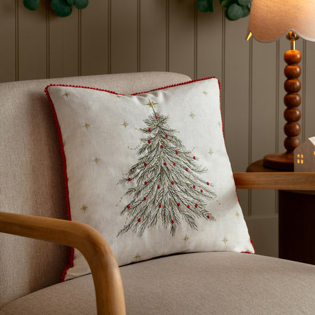 Twinkling Tree Velvet Linen Cushion in natural with red pom-pom trim, embroidered fir tree, gold stars and red baubles, styled on an armchair beside a wooden lamp.