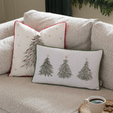 Rectangular cream velvet cushion with three embroidered green Christmas trees, red berries and gold stars, green pompom trim, shown on a sofa beside a red-trimmed cushion.