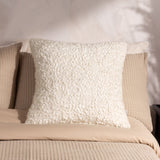 Ulsmere Wool Cotton Cushion in ecru with textured bouclé fabric, square shape, shown propped against pillows on a bed with beige linens.