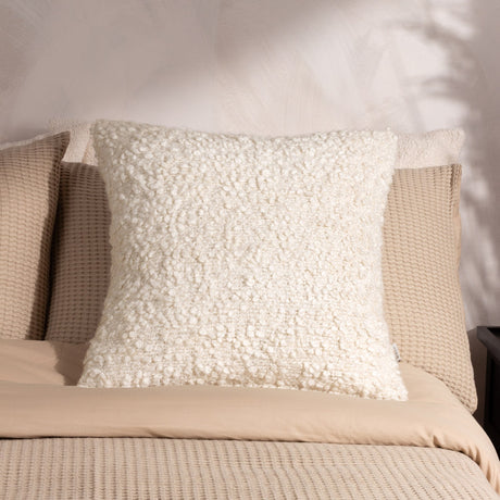 Ulsmere Wool Cotton Cushion in ecru with textured bouclé fabric, square shape, shown propped against pillows on a bed with beige linens.