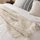 Ulsmere Wool Cotton Cushion in ecru with a bouclé, looped texture, styled on a bed alongside a striped cushion and a cream fringed throw.