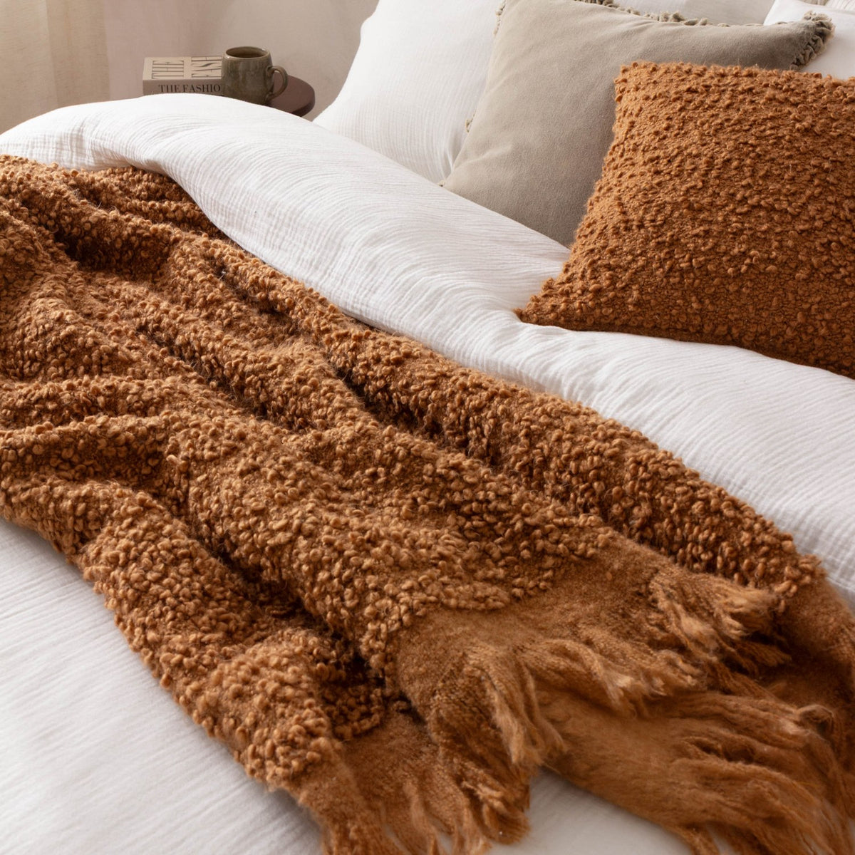 Ulsmere Wool Cotton Cushion in Ginger, a square boucle-textured cushion with dense looped orange-brown pile, styled on a white bedspread beside a matching throw.