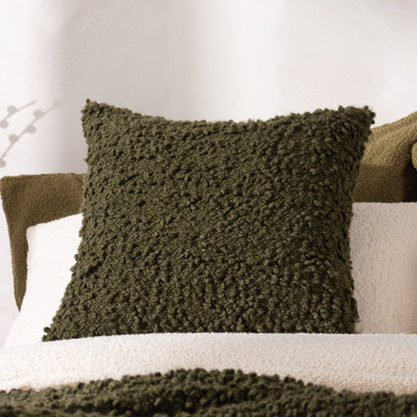 Ulsmere Wool Cotton Cushion in Lichen, a square textured bouclé cushion in dark olive green, shown on a bed with cream boucle bedding and a white sheer curtain backdrop.