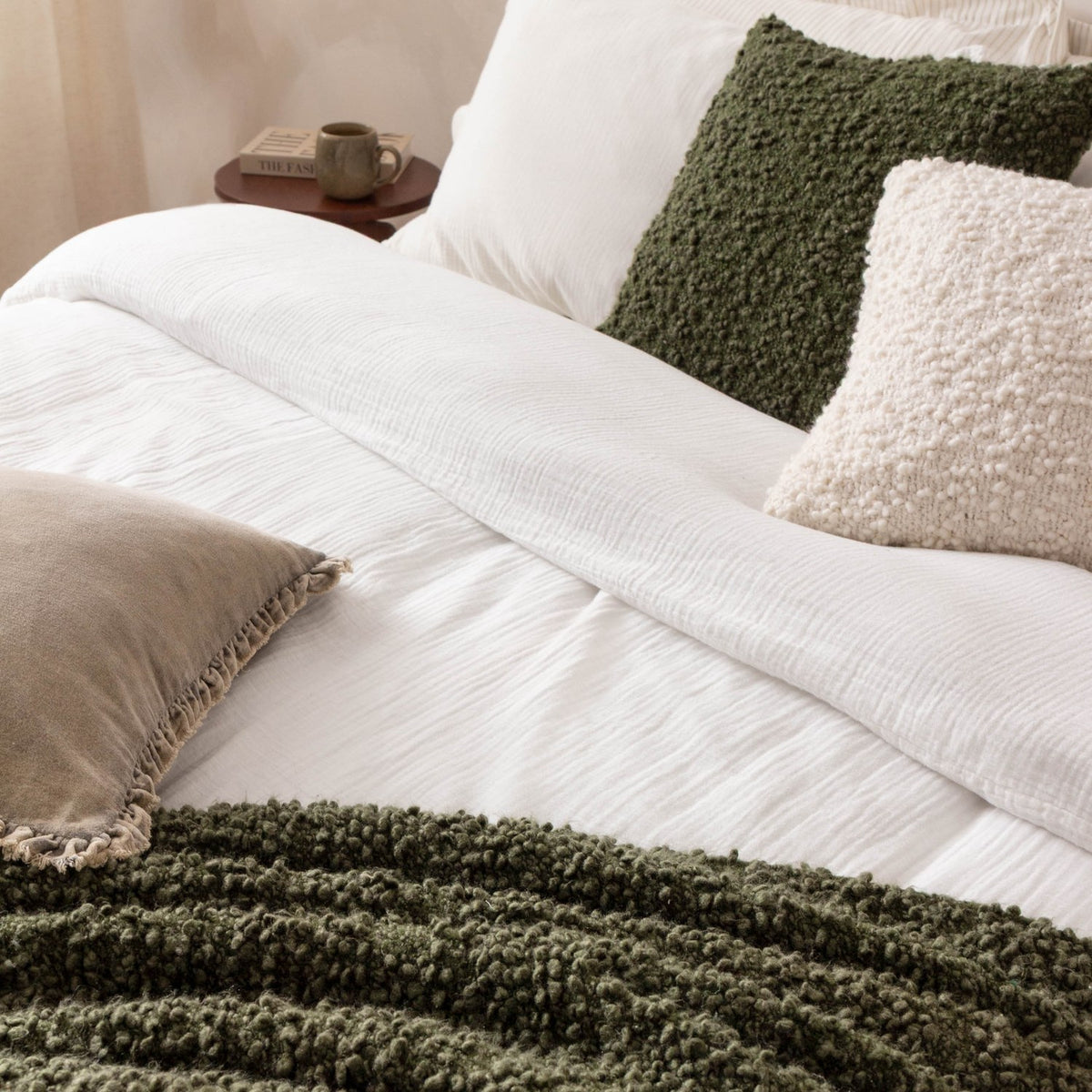 Ulsmere Wool Cotton Cushion in Lichen green bouclé, styled on a white bed with a cream boucle cushion and green throw in a bedroom setting.