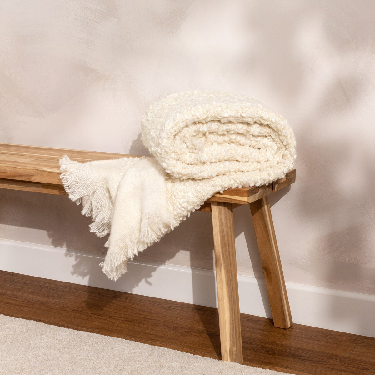 Ulsmere Reversible Wool Throw in ecru, folded with a fringed edge draped over a wooden bench, showing its bouclé texture against a neutral wall.