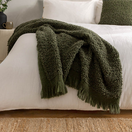 Ulsmere Reversible Wool Throw in Lichen green bouclé fabric with fringed edges, draped over a white bed in a bedroom setting.