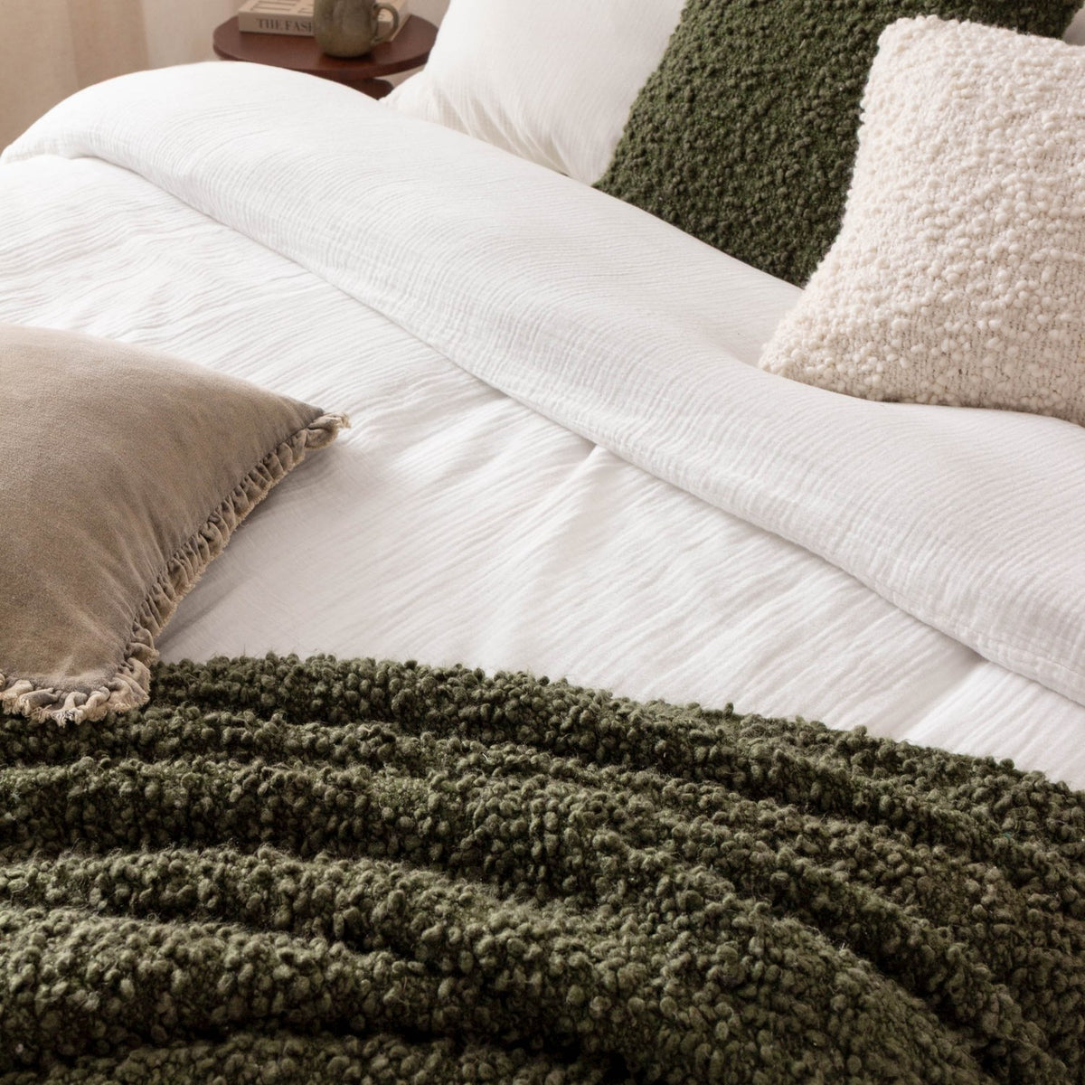 Ulsmere Reversible Wool Throw in Lichen green bouclé texture draped over a white bed with cream and olive cushions in a bedroom setting.