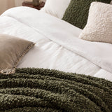 Ulsmere Reversible Wool Throw in Lichen green bouclé texture draped over a white bed with cream and olive cushions in a bedroom setting.