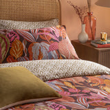Reversible floral duvet set in copper glow, with botanical leaf pillowcases in rust, pink and slate blue, olive diamond-print reverse, and mustard cushion, on a bed with a rattan headboard.