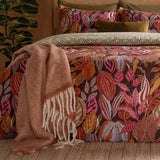Utopia Reversible Floral Duvet Cover Set in Copper Glow on a bed with botanical leaf print in rust, pink and slate blue on aubergine, styled with a rust fringed throw and olive cushions.