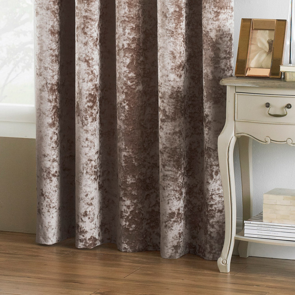 Verona Velvet Fully Lined Eyelet Curtains in Oyster, crushed velvet fabric with mottled brown-putty sheen, shown hanging beside a cream bedside table in a living room setting.