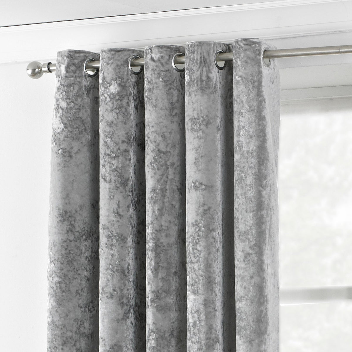 Verona silver crushed velvet eyelet curtain panel with stainless steel grommets hung on a metal pole, mottled fabric texture near a bright window.
