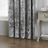 Verona Velvet Fully Lined Eyelet Curtains in silver, crushed velvet fabric with mottled texture, shown hanging beside a wooden side table on a hardwood floor.