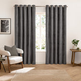 Verso Plain Reversible Eyelet Curtains in charcoal suede-effect fabric with grommet header, shown fully drawn either side of a window in a living room with armchair and side table.