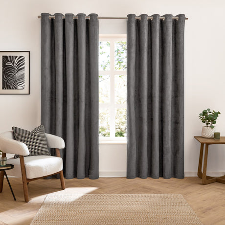 Verso Plain Reversible Eyelet Curtains in charcoal suede-effect fabric with grommet header, shown fully drawn either side of a window in a living room with armchair and side table.