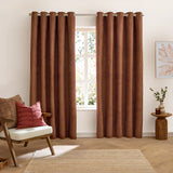 Verso Polyester Blackout Eyelet Curtains in cinnamon, a pair of velvety brown panels with eyelet heading, shown hanging in a living room beside a window.