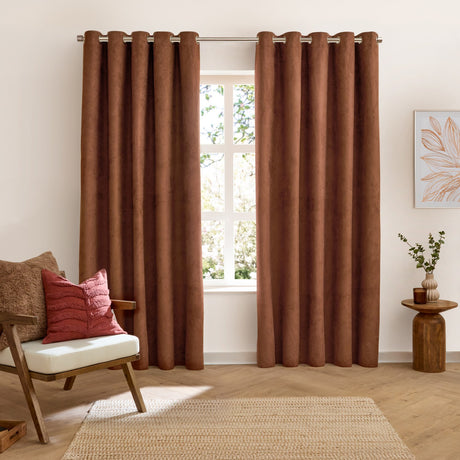 Verso Polyester Blackout Eyelet Curtains in cinnamon, a pair of velvety brown panels with eyelet heading, shown hanging in a living room beside a window.