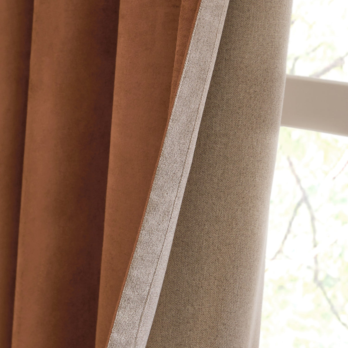 Close-up of Verso Polyester Blackout Eyelet Curtains in cinnamon, showing the velvety brown face and pale beige lining folded back near a window.
