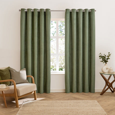 Verso Plain Reversible Eyelet Curtains in green suede-effect fabric with silver eyelets, shown floor-length either side of a window in a living room with an armchair and side table.
