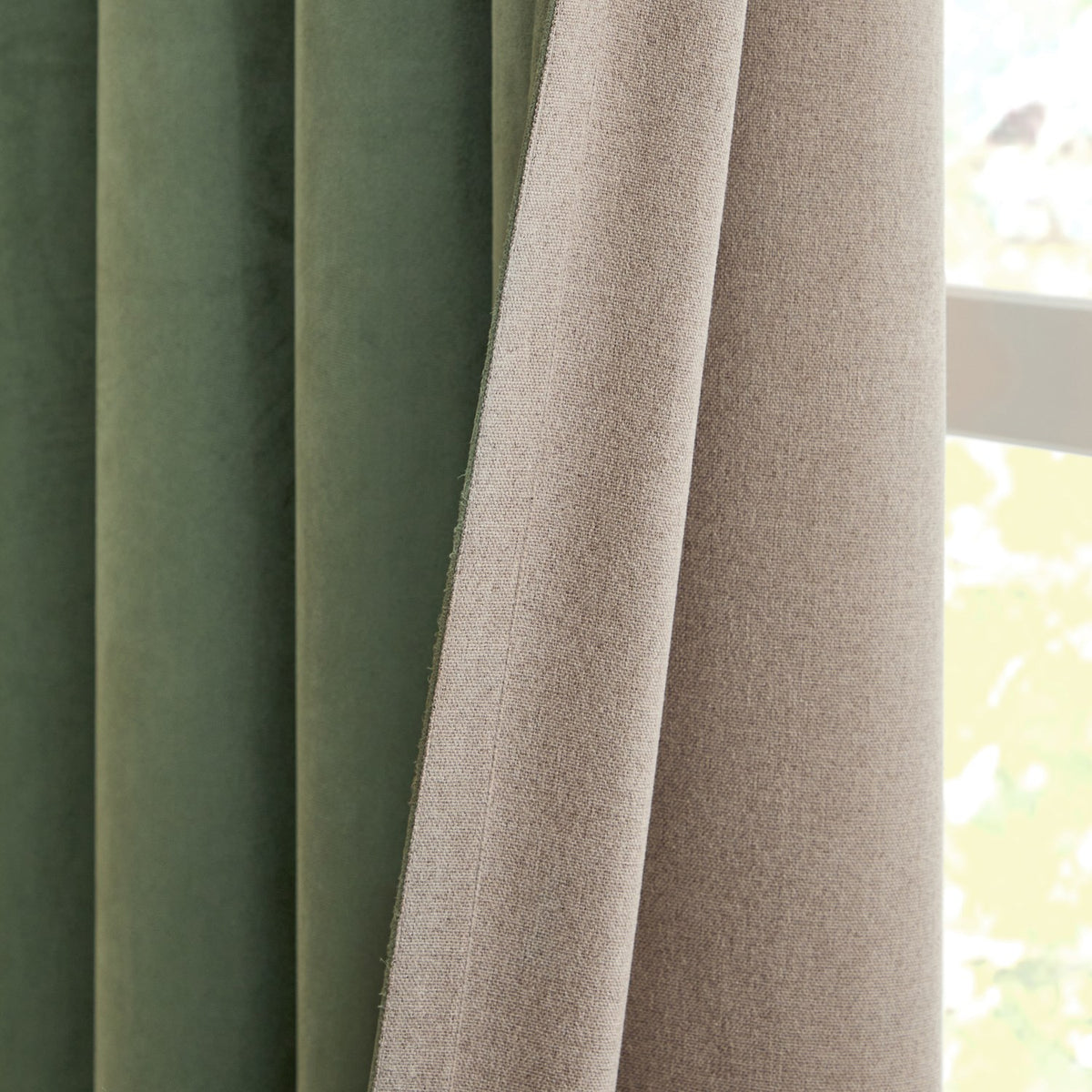 Verso Plain Reversible Eyelet Curtains in Green, close-up showing folded panels with green suede-effect fabric on one side and beige woven fabric on the reverse, beside a window.
