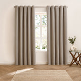 Verso Plain Reversible Eyelet Curtains in beige, hung either side of a window on a metal pole in a bedroom with wood flooring and a woven rug.