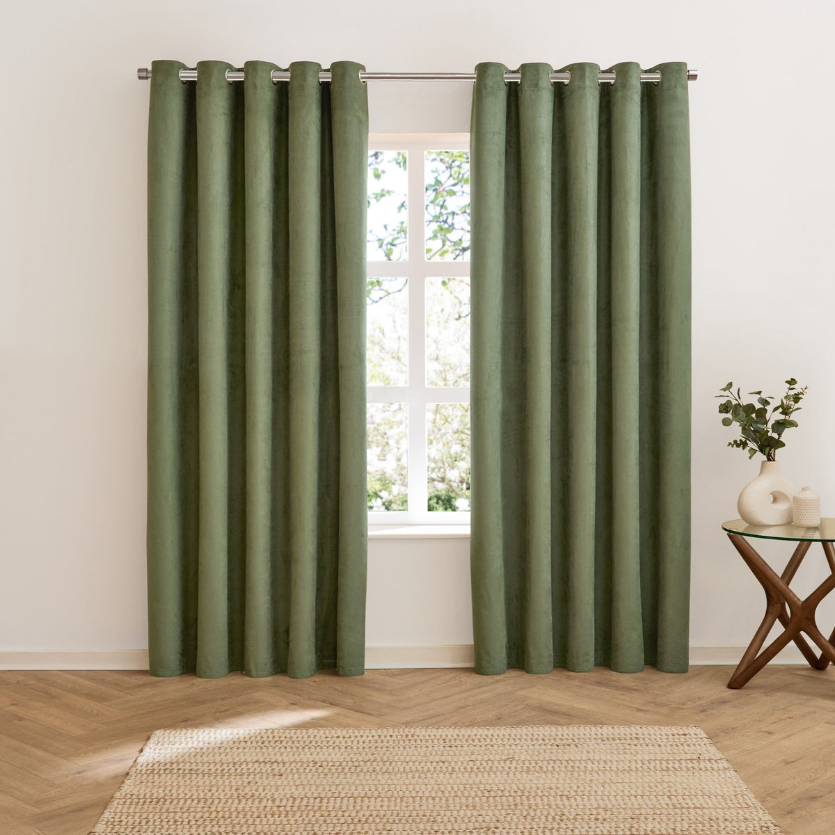 Verso Plain Reversible Eyelet Curtains in green suede-effect fabric hung on a metal rod either side of a window, shown in a living room with wood flooring and a jute rug.