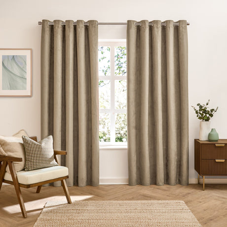 Verso Plain Reversible Eyelet Curtains in mocha, shown as full-length panels with silver eyelets on a pole, styled in a living room beside a wooden armchair and chest of drawers.