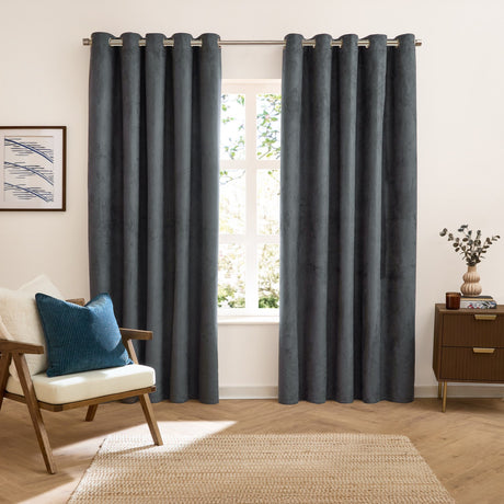 Verso Plain Reversible Eyelet Curtains in petrol blue with a suede-look finish, hung on a metal rail in a living room with an armchair, teal cushion and wooden sideboard.