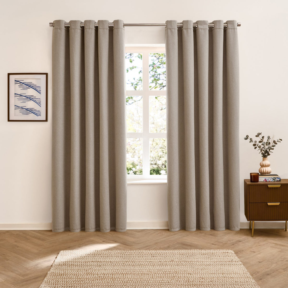 Verso Plain Reversible Eyelet Curtains in natural oatmeal shown floor-length either side of a window in a living room, styled with a wall print, sideboard and vase.
