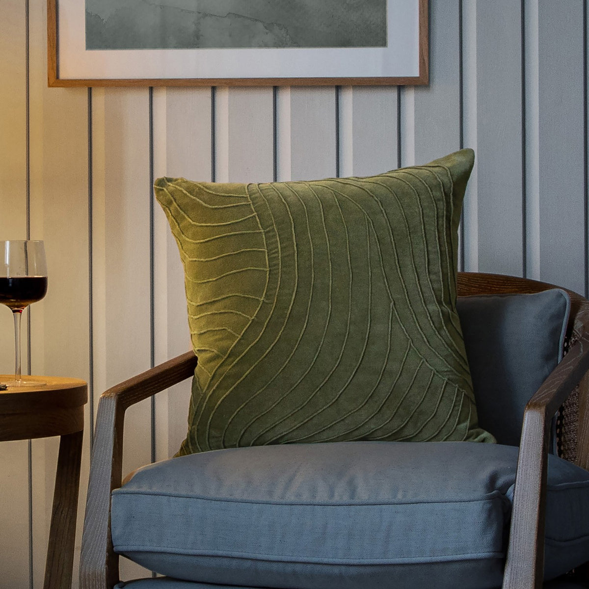 Waterfall Abstract Velvet Cushion in sage green with raised pintuck wave-like ridges, shown on a blue armchair in a living room setting.