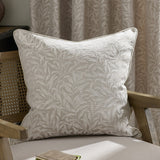 Square Willow Bough jacquard linen cushion with woven tonal leaf pattern in natural cream, piped edges, shown on a wooden armchair against curtains.
