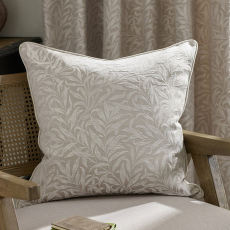 Square Willow Bough jacquard linen cushion with woven tonal leaf pattern in natural cream, piped edges, shown on a wooden armchair against curtains.