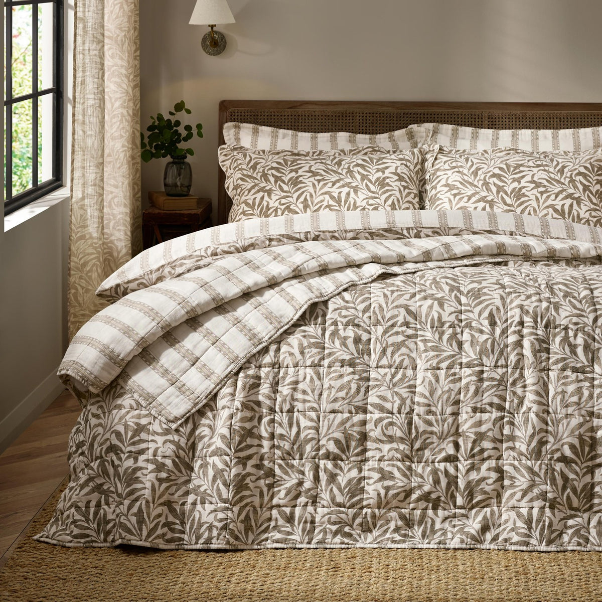 Willow Boughs reversible cotton quilted bedspread in taupe-brown leaf print on cream, styled on a bed with matching striped pillow shams in a neutral bedroom.