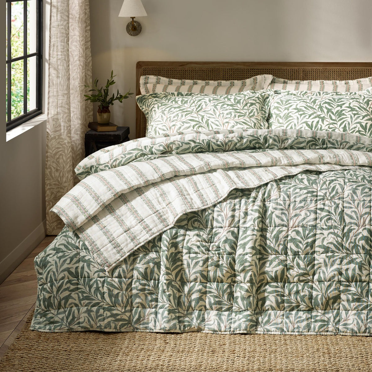 Willow Boughs reversible cotton bedspread in sage, quilted with a trailing leaf print, shown on a bed with matching pillow shams and a striped reverse in a bedroom setting.