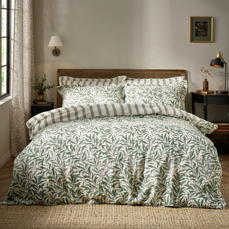 Willow Boughs cotton duvet cover set in sage green willow leaf print with striped-reverse pillowcases, shown on a bed with cane headboard in a bedroom setting.