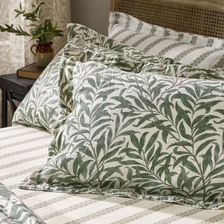 Willow Boughs reversible cotton pillowcase pair in sage, styled on a bed with green willow leaf print pillowcases against striped bedding in a bedroom setting.
