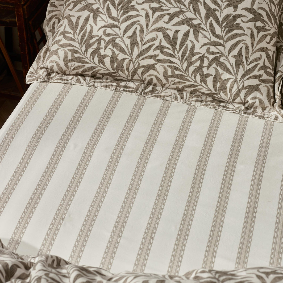 Willow Boughs Percale Cotton Fitted Sheet in Natural, showing a fine vertical stripe with dot detail in soft natural tones on white cotton, styled on a bed with matching leaf-print pillows.