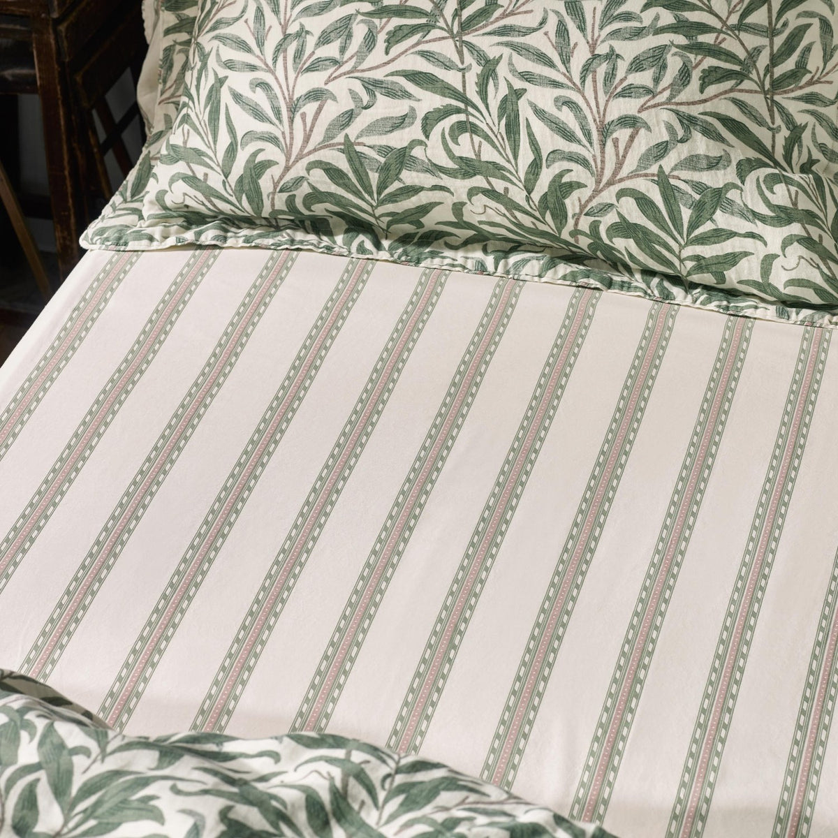Willow Boughs Percale Cotton Fitted Sheet in Sage, cream ground with fine green and pink striped pattern, shown on a bed with a matching Willow Boughs leaf-print pillowcase.