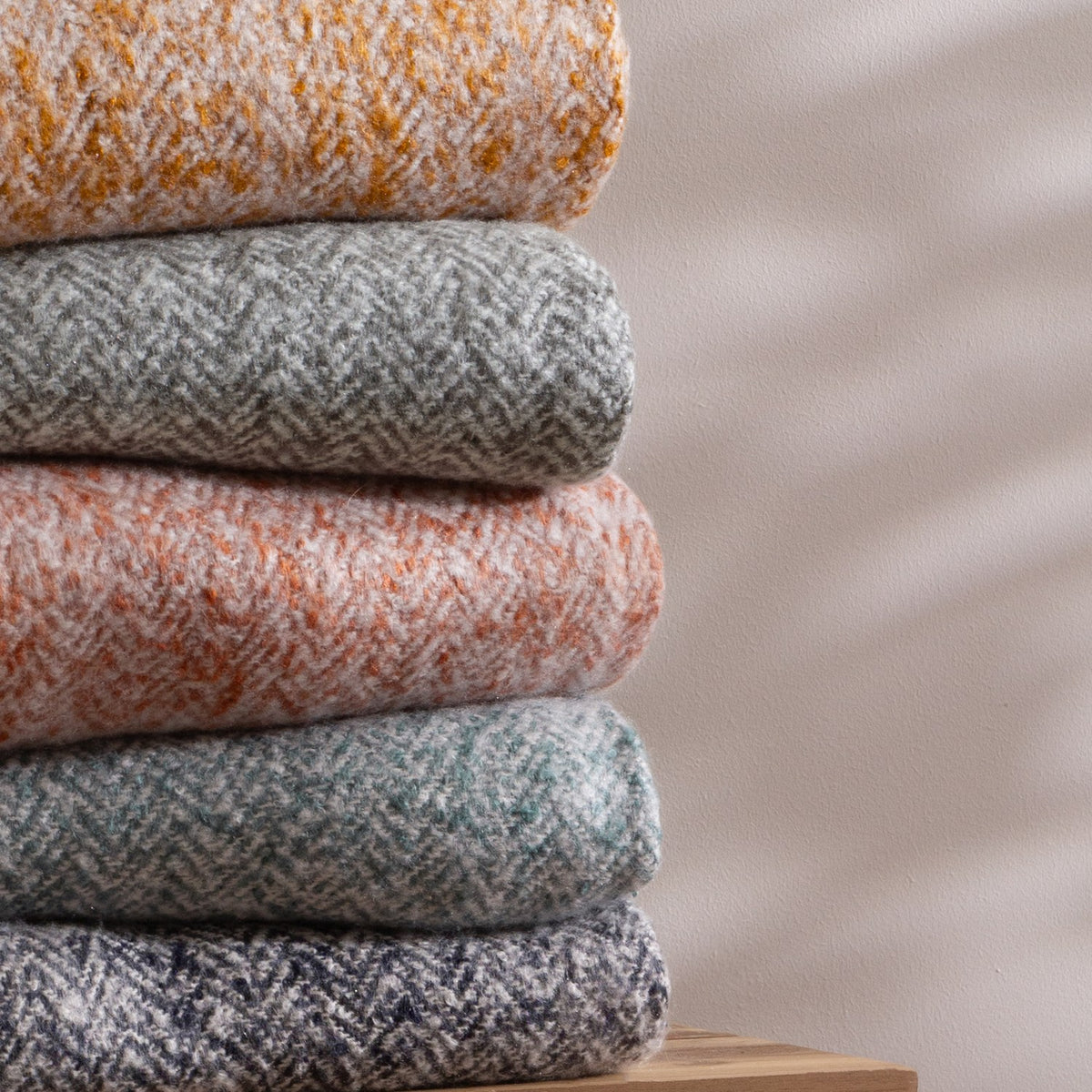 Weaver Geometric Wool Throw in gold, folded with herringbone weave in amber and cream tones, shown stacked with other coloured throws against a plain light background.