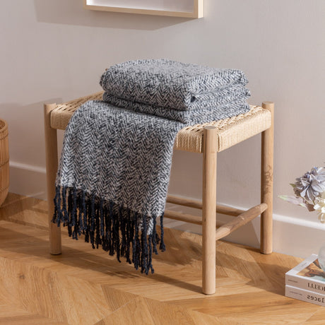 Weaver Geometric Wool Throw in navy herringbone weave, folded and draped with fringed edge over a wooden bench with woven rope seat in a light-toned room.