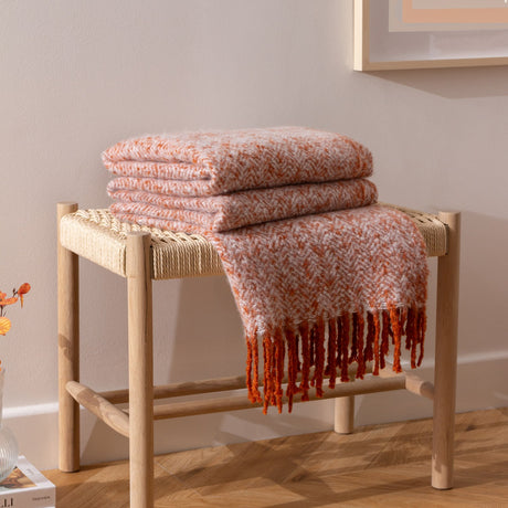 Weaver Geometric Wool Throw in rust, folded on a woven-seat wooden bench with fringed edge draping down, showing herringbone texture in rust and cream tones.