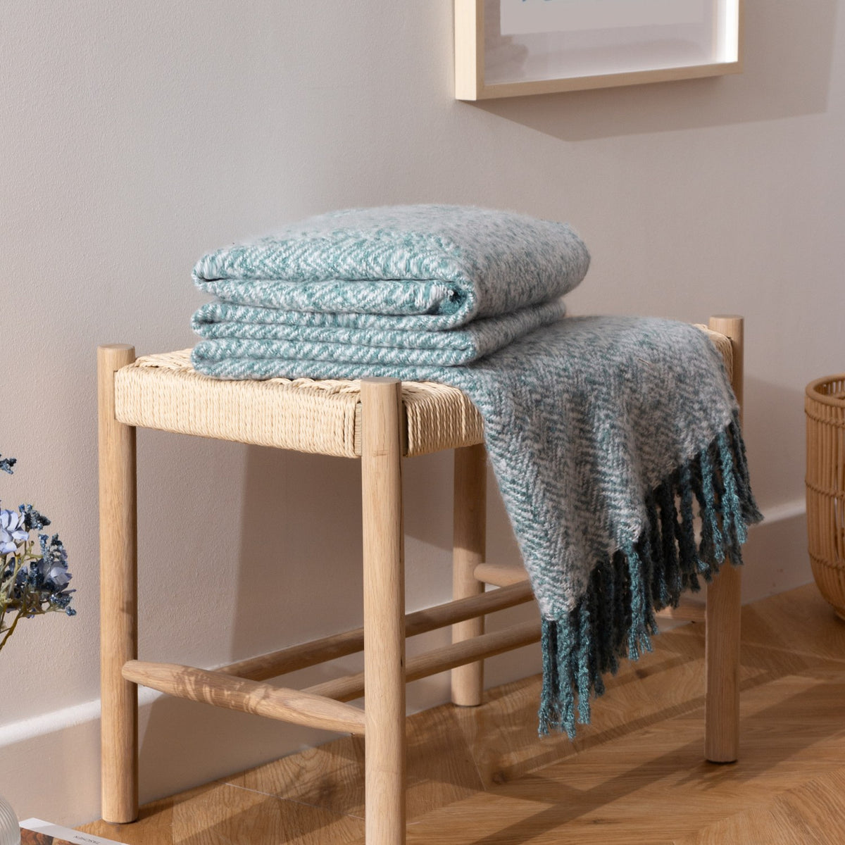 Weaver Geometric Wool Throw in teal herringbone weave, folded on a wooden bench with one length draped and tasselled edge visible, in a light-toned room.