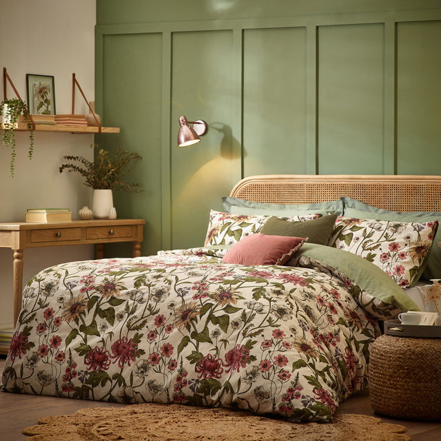 Wallflower Cotton Botanical Duvet Cover Set in Natural styled on a bed with rattan headboard, cream ground floral print, and green and pink cushions in a green-panelled bedroom.