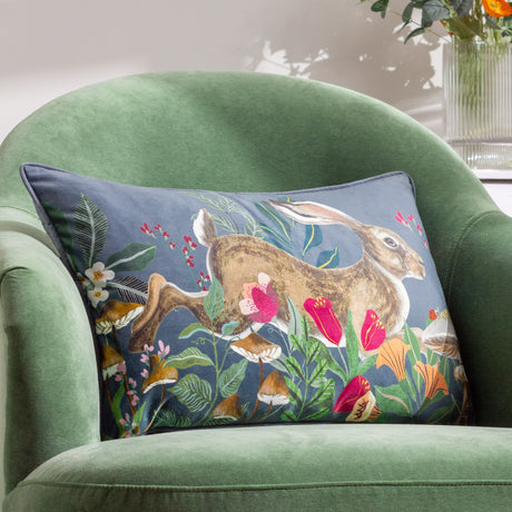 Rectangular velvet cushion in grey-blue with a printed leaping hare among poppies, ferns and toadstools, shown on a green armchair.