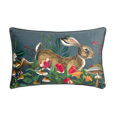 Rectangular blue-grey velvet cushion with piped edge, featuring a printed leaping hare among poppies, ferns and toadstools with a ladybird detail, shown on a white background.