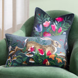 Wild Garden Animal Velvet Cushion in grey-blue with a hand-painted leaping hare among poppies and toadstools, styled on a green armchair with a floral navy cushion behind.