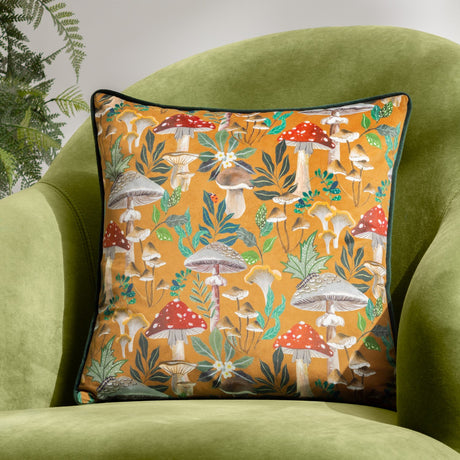Square velvet-fronted cushion from the Wildgarden Mushroom range, amber-gold ground with red-and-white toadstools, ferns and leaves, dark green piped edge, shown on a green armchair.